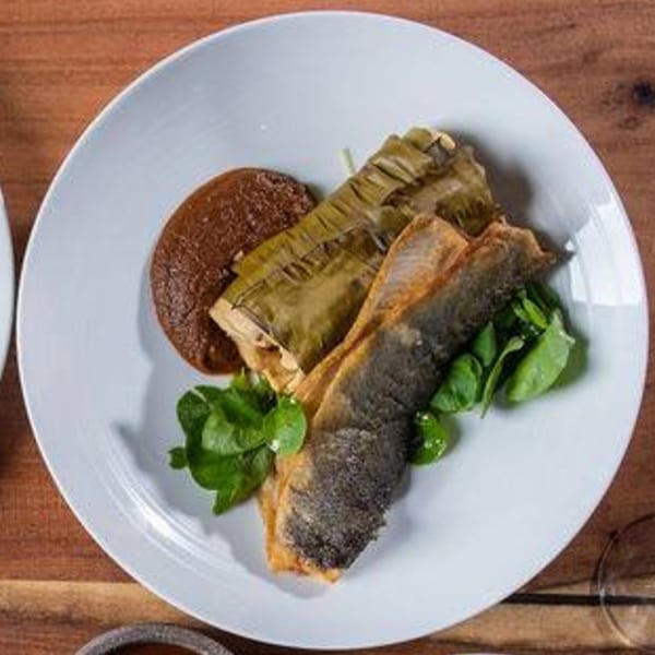 Ofrenda's Mexican dish with fish, tamale, dark sauce, and greens on a white plate.